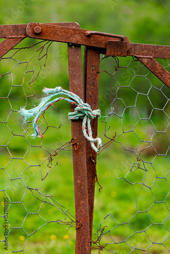A rusty gate made of metal fencing is tied with a blue rope, situated in a vibrant green field abundant with wildflowers.