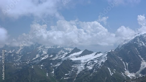 Camera Pans From Left To Right Across A Majestic Mountain Range With Snow-Capped Peaks And Glaciers Under A Blue Sky With Clouds. Scenic Alpine Landscape.