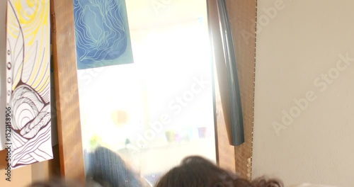 A young woman examines her reflection in a large mirror while holding a cozy sweater. The walls are decorated with vibrant drawings, creating a warm, inviting atmosphere for introspection