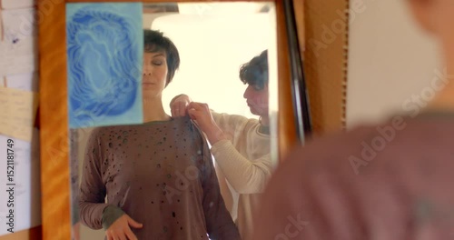 A fashion model, aided by a stylist, wears a trendy sweater in front of a mirror, showcasing teamwork and a shared passion for style and personal expression through clothing