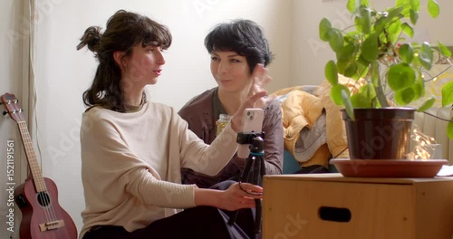 Two close friends are having fun engaging in filming with a smartphone, exchanging creative ideas in a warm and inviting indoor environment decorated with potted plants and casual decor