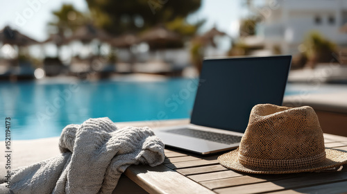 Relaxing laptop setup beside an inviting pool with a straw hat and towel on a sunny day in a tranquil resort environment
