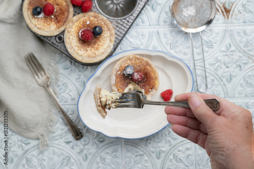 Hand holding fork with a bite of cream cheese tartlet or mini pie with cacao espresso powder and fresh raspberries and blueberries
