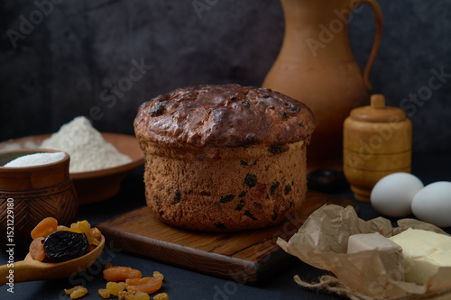 A round loaf of easter cake sits on a wooden board, surrounded by eggs, butter, flour, sugar, yeast and dried fruits photo