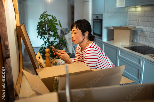 Asian woman using smartphone while unpacking boxes in her new home kitchen photo