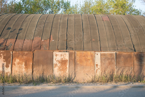 Old industrial building with rusted exterior and overgrown grass near a quiet road during sunset in a rural area