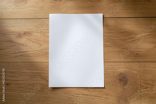 Top view of a verticle blank white A4 paper laying on a medium oak with natural window light, for product photography. 
