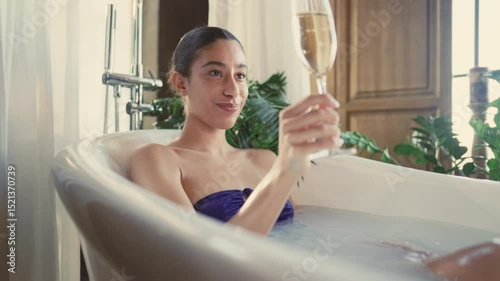 Happy young woman taking bath, drinking champagne alone, celebrating birthday. Beautiful mixed race lady relaxing and enjoying sparkling wine, single party in bathroom in weekend, enjoy solitude