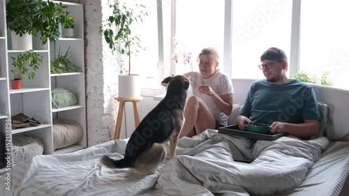 happy young millennial couple having breakfast in bed and enjoying fun time with their dog