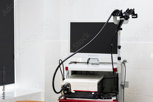 A modern endoscopy system with a large monitor, processing unit, and endoscopes is on a mobile cart in a clean medical room with tiled walls.