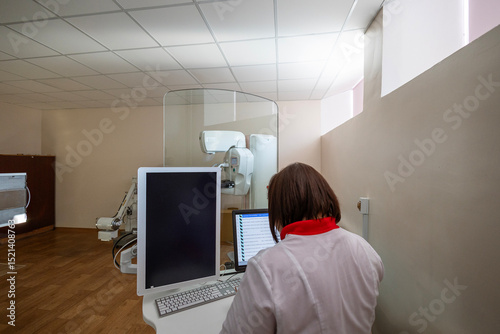 A technician operates a mammography machine from a control station with dual monitors, separated by a transparent partition in a medical room.