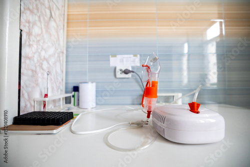 A close-up view shows a person's hands holding an inhaler device with an orange mask. The device is connected to a white compressor, all resting on a white table in a medical setting with tiled walls