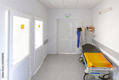A sterile hospital admission department corridor features a modern stretcher, a mobile X-ray machine, and bright white doors, including an exit.