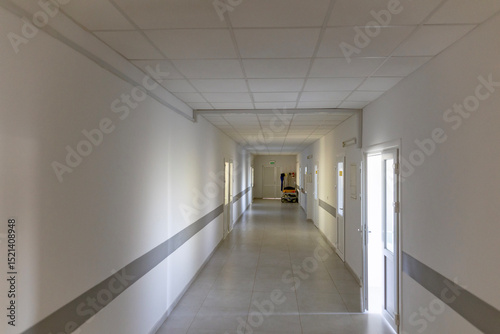 A long, empty hospital corridor with white walls, a grey stripe, and multiple doors. The sterile hallway features a tiled floor, and at the end of the corridor, a hospital bed with a catheter stand