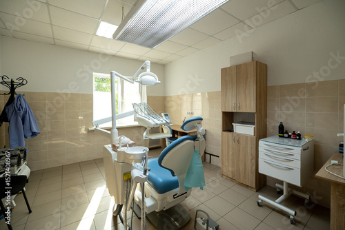 A modern dental clinic room features a blue dental chair, white dental unit with instruments, overhead operating light, and wooden cabinetry in a clean treatment space.