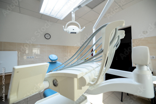 A modern dental clinic room features a blue dental chair, white dental unit with instruments, an overhead operating light, and a wall clock in a clean setting.