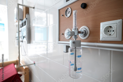 An oxygen hub with a flow meter and humidifier bottle is mounted on a wooden-paneled wall above a hospital bed in an intensive care unit