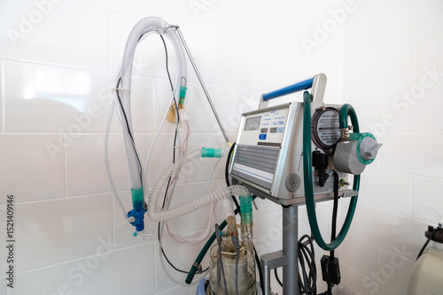 A ventilator, a complex medical apparatus with tubing and controls, stands next to a hospital bed in a clean, tiled room