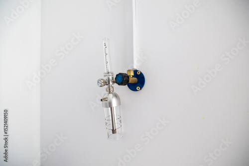An oxygen flow meter with a humidifier bottle is mounted on a white wall, part of an oxygen hub, ready for use in a hospital intensive care unit.