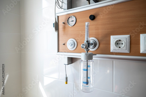 An oxygen hub with a flow meter and humidifier bottle is mounted on a wooden-paneled wall above a hospital bed in an intensive care unit