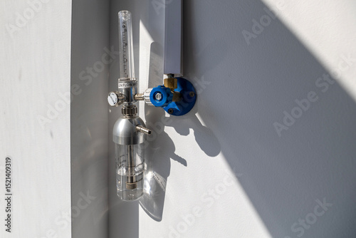An oxygen flow meter with a humidifier bottle is mounted on a white wall, part of an oxygen hub, ready for use in a hospital intensive care unit.