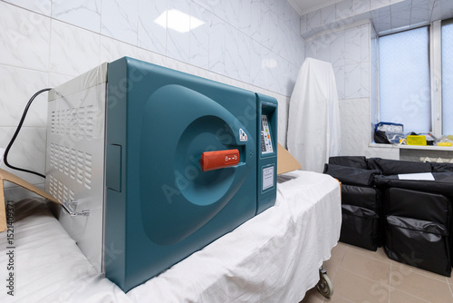 A new, unplugged medical sterilizer, an autoclave, rests on a gurney in a clean hospital room with tiled walls and nearby equipment bags.