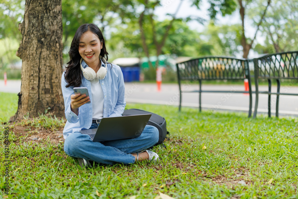 Happy asian freelancer working remotely outdoors using laptop and smartphone, enjoying advantages of freelance work and flexible schedule, sitting on grass under tree in city park Happy asian freelancer working remotely outdoors using laptop and smartphone, enjoying advantages of freelance work and flexible schedule, sitting on grass under tree in city park