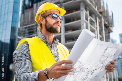 Builder in yellow vest and hard hat inspects blueprint against background of unfinished building