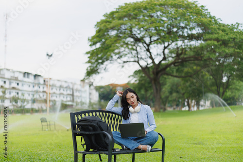 Concentrated young female student is working on laptop while sitting on bench in park with backpack near her, enjoying good weather and fresh air
