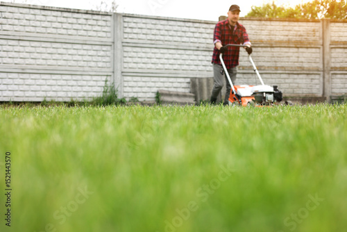 view of man in casual clothes mows lawn with lawn mower at backyard of his house. Husband takes care of garden on spring cloudy day. Modern gasoline garden equipment. Landscaping work