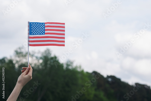 Close-up of a female hand holding USA flag against a natural outdoor background with cloudy sky and green trees. Concept of patriotism, national pride, and Independence Day celebration