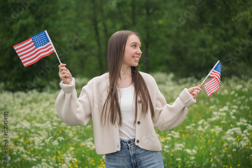 Young woman smiling and holding two USA flags in lush green field with wildflowers on 4th of July. Concept of patriotism, national pride and Independence Day celebration
