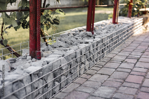 Stone fence in steel mesh and green lawn. Close up of gabion with tightly packed stones of varying sizes and textures. Combination of natural materials and industrial design.