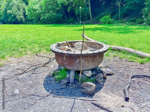 Rusty metal fire pit with stones and sticks in a sunlit park clearing, surrounded by trees and open grassy field.