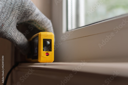 A gloved hand holds a yellow laser distance meter against a window sill, focusing on precise measurement in home renovation or construction projects.