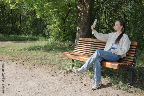 Young woman sitting on a bench in a sunny park, taking selfie with smartphone. Concept of digital communication, leisure, outdoor lifestyle, springtime relaxation and solo travel.