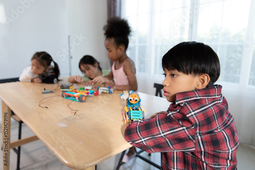 A young boy thoughtfully observes his robotics project, as other children in the background engage in a hands-on STEM learning activity with colorful construction toys.