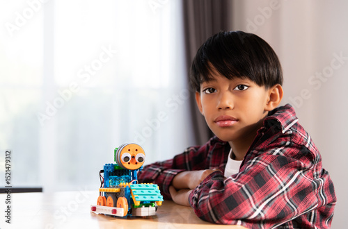A young boy, likely of East Asian descent, looks directly at the camera with a serious expression, sitting beside a colorful, self-assembled robot.