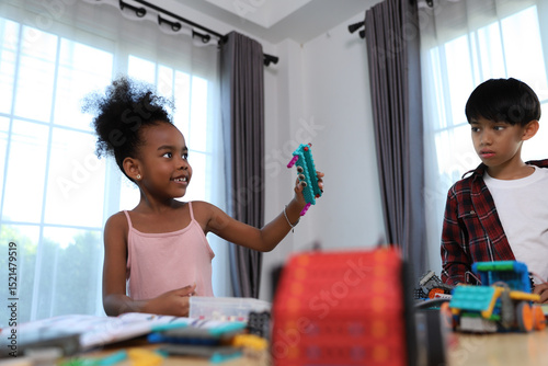A joyful young girl proudly holds up her colorful construction toy creation, sharing her excitement with a curious boy during a fun and engaging STEM activity.
