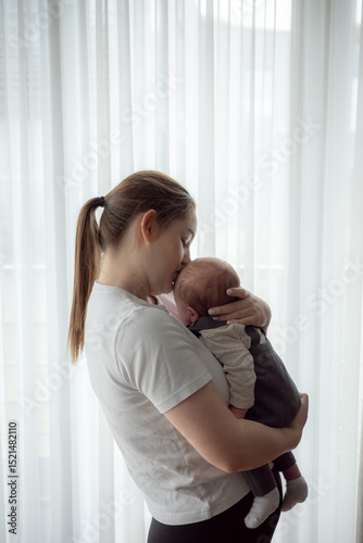Young mother holding newborn baby in soft natural light standing by a window at home. Concept of early motherhood, tenderness, bonding, parental love, and infant care in a calm home environment.