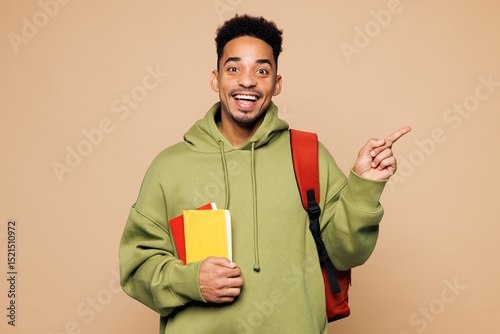 Young happy boy student he wear green casual clothes backpack bag hold books point index finger aside on area isolated on plain pastel light beige background. High school university college concept.