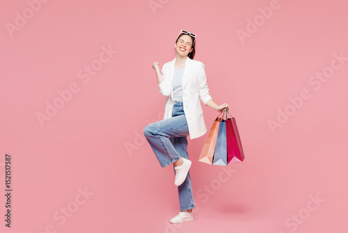 Full body young woman wear white clothes hold shopping paper package bags do winner gesture celebrate clenching fists say yes isolated on plain pink color background Black Friday sale buy day concept