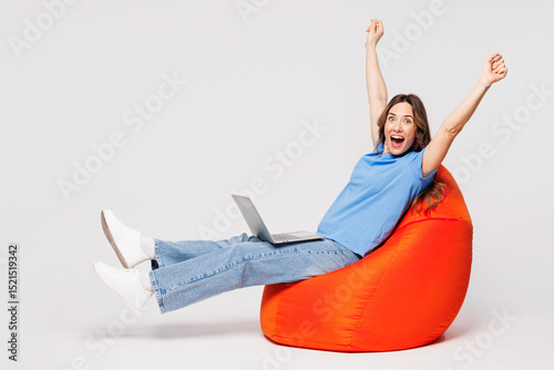 Full body young IT woman she wears blue t-shirt casual clothes sit in bag chair hold use work on laptop pc computer do winner gesture isolated on plain solid white color background. Lifestyle concept.