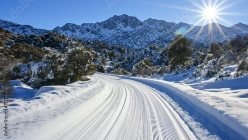 Curved snow-covered road winding through pine forest toward alpine mountain under clear blue sky and sunburst. Transportation concept