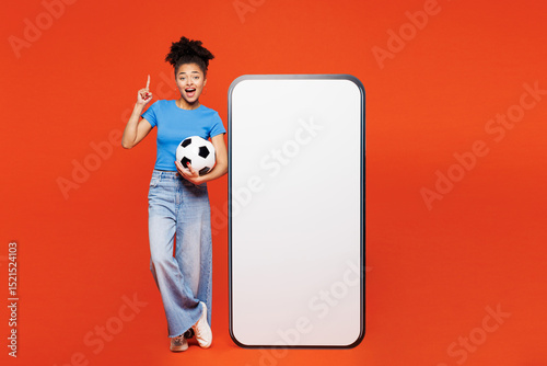 Full body young woman fan in blue clothes support football sport team hold soccer ball watch tv live stream big blank screen mobile cell phone point finger up isolated on plain red orange background