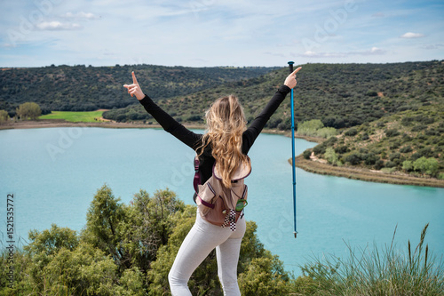 Teen girl pointing skyward with trekking pole while enjoying panoramic view of turquoise lagoon from scenic hillside during spring hike