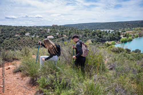 Teen girl and boy hiking through rugged hillside terrain, using walking poles to navigate a natural trail with a turquoise lagoon and village in the background