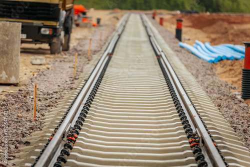 Newly laid railway track under construction, surrounded by building materials, piping, and vehicles. Construction equipment and supplies are visible along the gravel base.