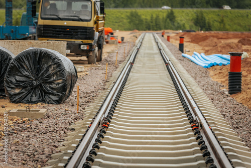 Newly laid railway track under construction, surrounded by building materials, piping, and vehicles. Construction equipment and supplies are visible along the gravel base.