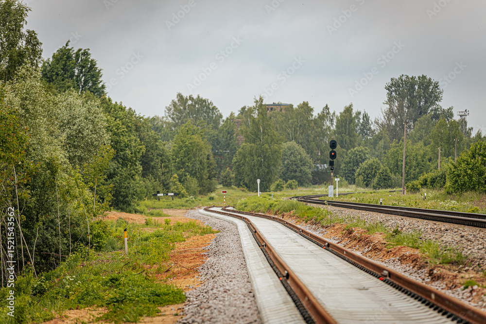 Obraz premium A curved section of newly laid railway track diverges beside an older line, with a circular signal sign and red light signal post in a green, forested area.